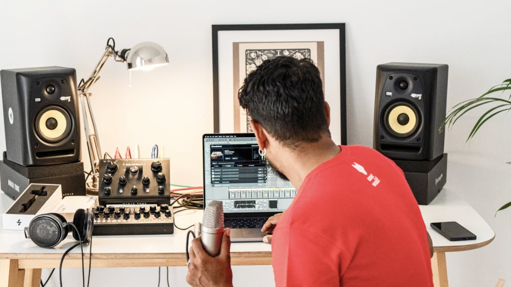 A music producer in a red shirt holds a microphone while working on a laptop in a home studio, surrounded by studio monitors, audio equipment, and headphones.